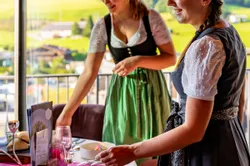 Zwei Frauen in Dirndl kleiden einen elegant gedeckten Tisch mit Alpenblick